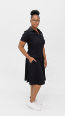A woman modeling a black short-sleeve collared dress with side pockets, paired with white sneakers, standing against a seamless white studio background.