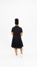 A woman viewed from behind wearing a black short-sleeve collared dress with a flared skirt and white sneakers, standing against a seamless white studio background.