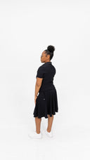 A woman wearing a black short-sleeve collared top and a black pleated midi skirt, paired with white sneakers, standing in profile against a solid white studio background.